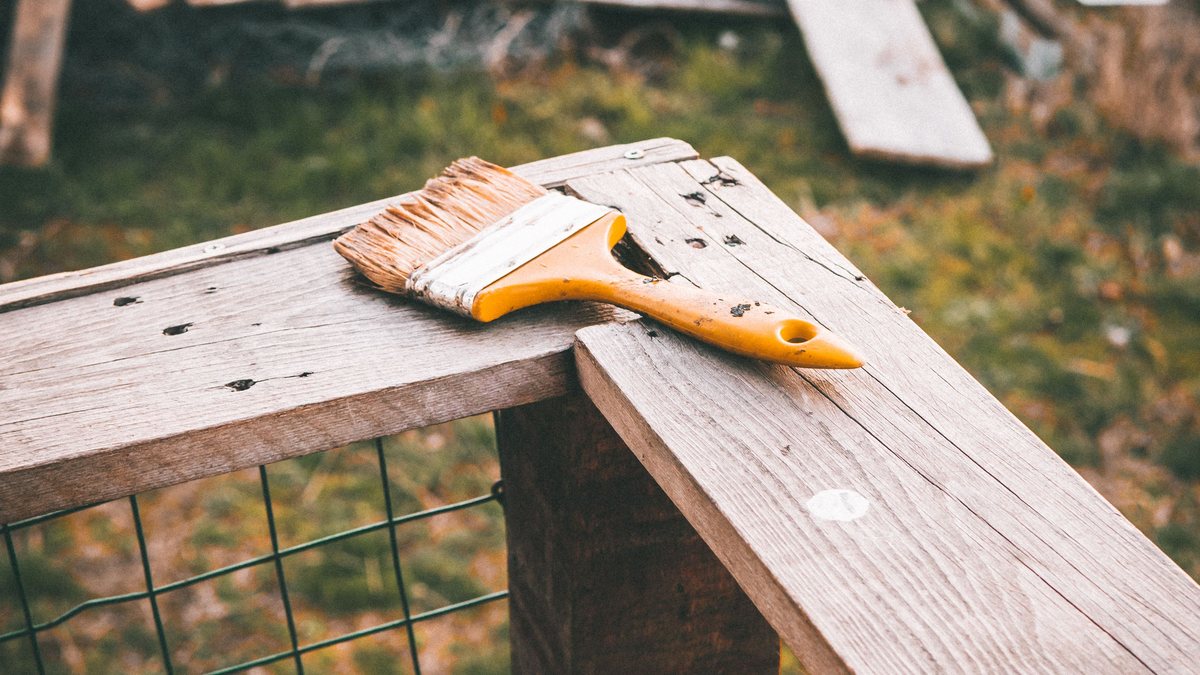 Yellow-handled paintbrush resting across weathered outdoor wooden boards before maintenance.